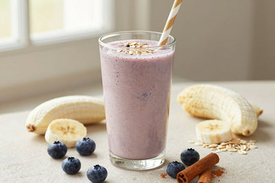 A tall glass of thick, creamy blueberry muffin protein shake with a straw, a few of its fresh ingredients beside it