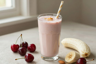 A tall glass of thick, creamy cherry almond protein shake with a straw, a few of its fresh ingredients beside it