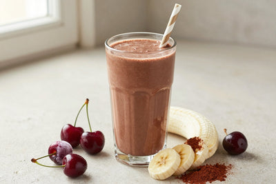 A tall glass of thick, creamy chocolate cherry protein shake with a straw, a few of its fresh ingredients beside it