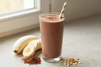A tall glass of thick, creamy chocolate oat protein shake with a straw, a few of its fresh ingredients beside it