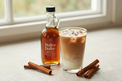 A small glass bottle of cinnamon dolce syrup with a pour spout, beside an iced latte sweetened with it