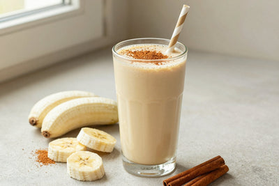A tall glass of thick, creamy cinnamon roll protein shake with a straw, a few of its fresh ingredients beside it
