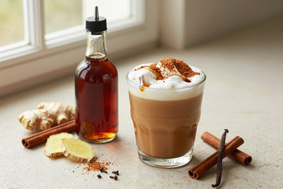 A small glass bottle of gingerbread syrup with a pour spout, beside an iced latte sweetened with it