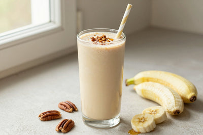 A tall glass of thick, creamy maple pecan protein shake with a straw, a few of its fresh ingredients beside it