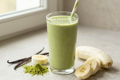 A tall glass of thick, creamy matcha vanilla protein shake with a straw, a few of its fresh ingredients beside it