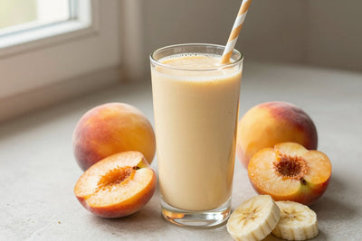 A tall glass of thick, creamy peach protein shake with a straw, a few of its fresh ingredients beside it
