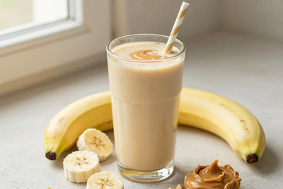 A tall glass of thick, creamy peanut butter banana protein shake with a straw, a few of its fresh ingredients beside it