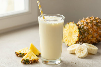 A tall glass of thick, creamy pineapple greek yogurt protein shake with a straw, a few of its fresh ingredients beside it