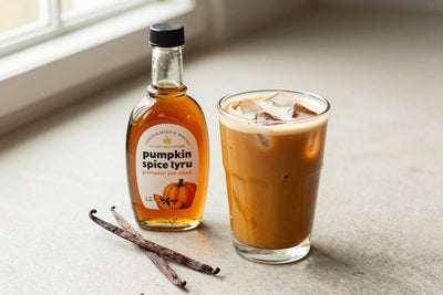 A small glass bottle of pumpkin spice syrup with a pour spout, beside an iced latte sweetened with it