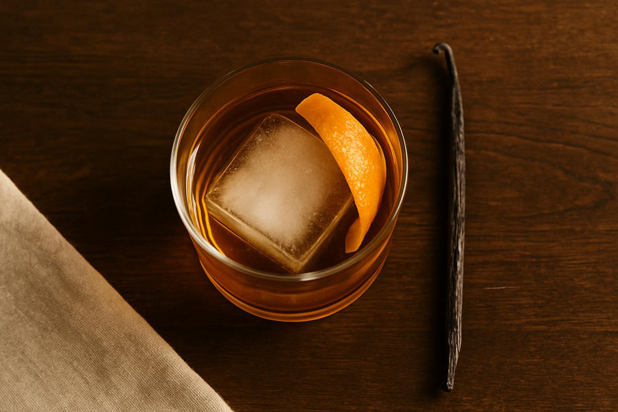 Vanilla old fashioned cocktail with large ice cube and orange peel garnish on dark wood bar