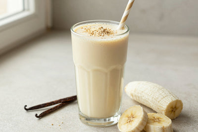 A tall glass of thick, creamy vegan vanilla protein shake with a straw, a few of its fresh ingredients beside it