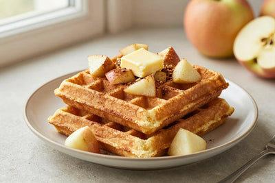 A stack of golden apple cinnamon waffles on a plate with a pat of butter and the toppings from the recipe