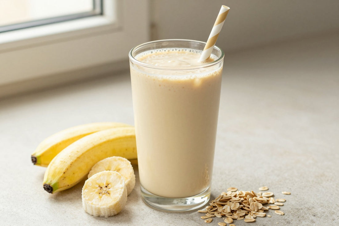 A tall glass of thick, creamy banana oat protein shake with a straw, a few of its fresh ingredients beside it