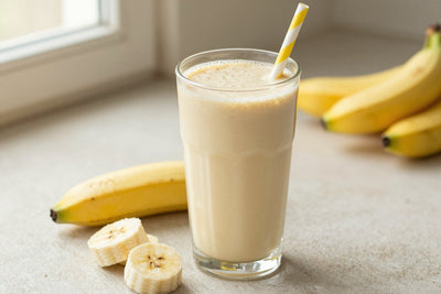 A tall glass of thick, creamy banana protein shake with a straw, a few of its fresh ingredients beside it