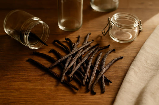Scattered Madagascar Grade B vanilla beans on a wooden kitchen table next to open glass jars, overhead angle, warm natural