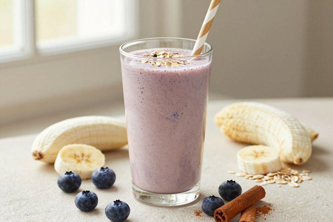 A tall glass of thick, creamy blueberry muffin protein shake with a straw, a few of its fresh ingredients beside it