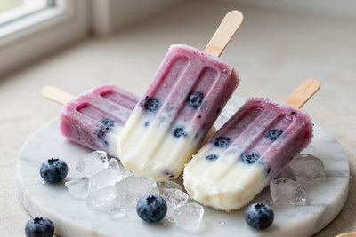 Three blueberry popsicles on a bed of crushed ice on a light marble surface, one with a bite taken out