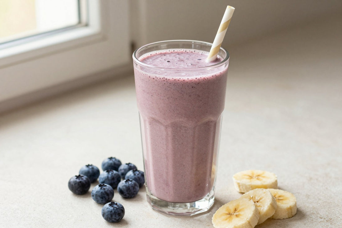 A tall glass of thick, creamy blueberry protein shake with a straw, a few of its fresh ingredients beside it