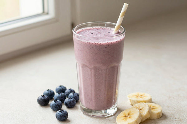 A tall glass of thick, creamy blueberry protein shake with a straw, a few of its fresh ingredients beside it