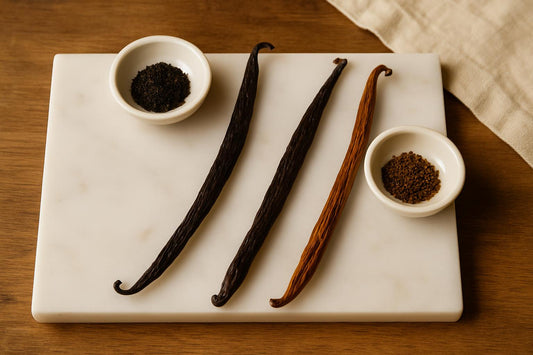 Three vanilla beans of different origins arranged on a marble surface with small white bowls containing vanilla caviar seeds