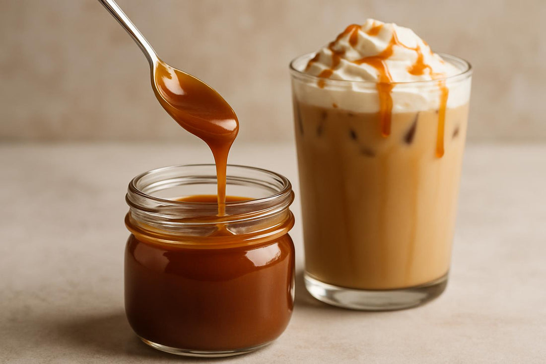 A small glass jar of glossy amber caramel sauce with a spoon lifting a ribbon of it, beside an iced caramel latte with carame