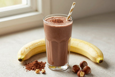 A tall glass of thick, creamy chocolate hazelnut protein shake with a straw, a few of its fresh ingredients beside it