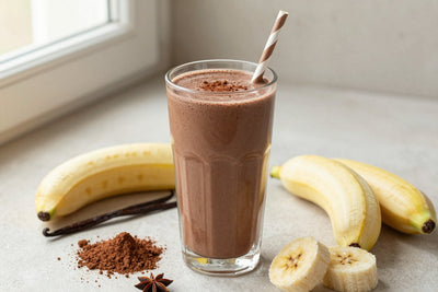 A tall glass of thick, creamy chocolate protein shake with a straw, a few of its fresh ingredients beside it
