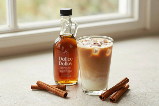 A small glass bottle of cinnamon dolce syrup with a pour spout, beside an iced latte sweetened with it