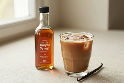 A small glass bottle of classic simple syrup with a pour spout, beside an iced latte sweetened with it