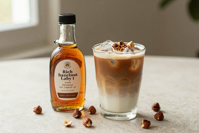 A small glass bottle of hazelnut syrup with a pour spout, beside an iced latte sweetened with it