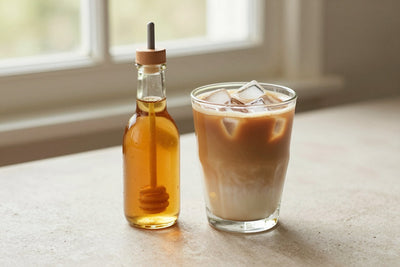 A small glass bottle of honey syrup with a pour spout, beside an iced latte sweetened with it