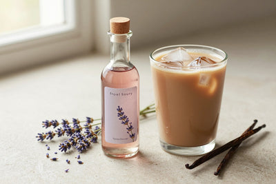 A small glass bottle of lavender syrup with a pour spout, beside an iced latte sweetened with it
