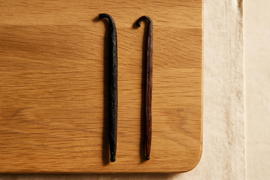 Two whole vanilla beans side by side on a light wooden cutting board, one Madagascar bean and one Mexican bean showing color