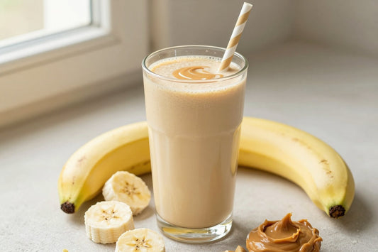 A tall glass of thick, creamy peanut butter banana protein shake with a straw, a few of its fresh ingredients beside it