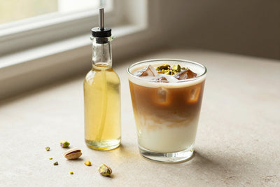 A small glass bottle of pistachio syrup with a pour spout, beside an iced latte sweetened with it