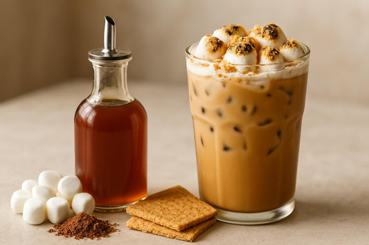 A small glass bottle of light brown s'mores syrup with a pour spout, beside an iced latte topped with toasted marshmallows an