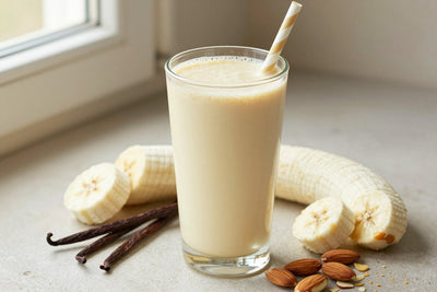 A tall glass of thick, creamy vanilla almond protein shake with a straw, a few of its fresh ingredients beside it