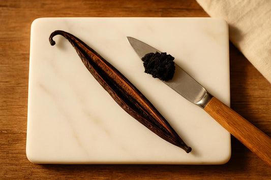Split Madagascar vanilla bean on white marble surface with heap of scraped black caviar seeds on knife blade beside empty