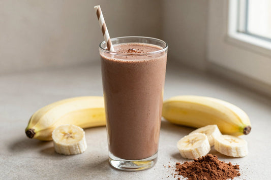 A tall glass of thick, creamy vegan chocolate protein shake with a straw, a few of its fresh ingredients beside it