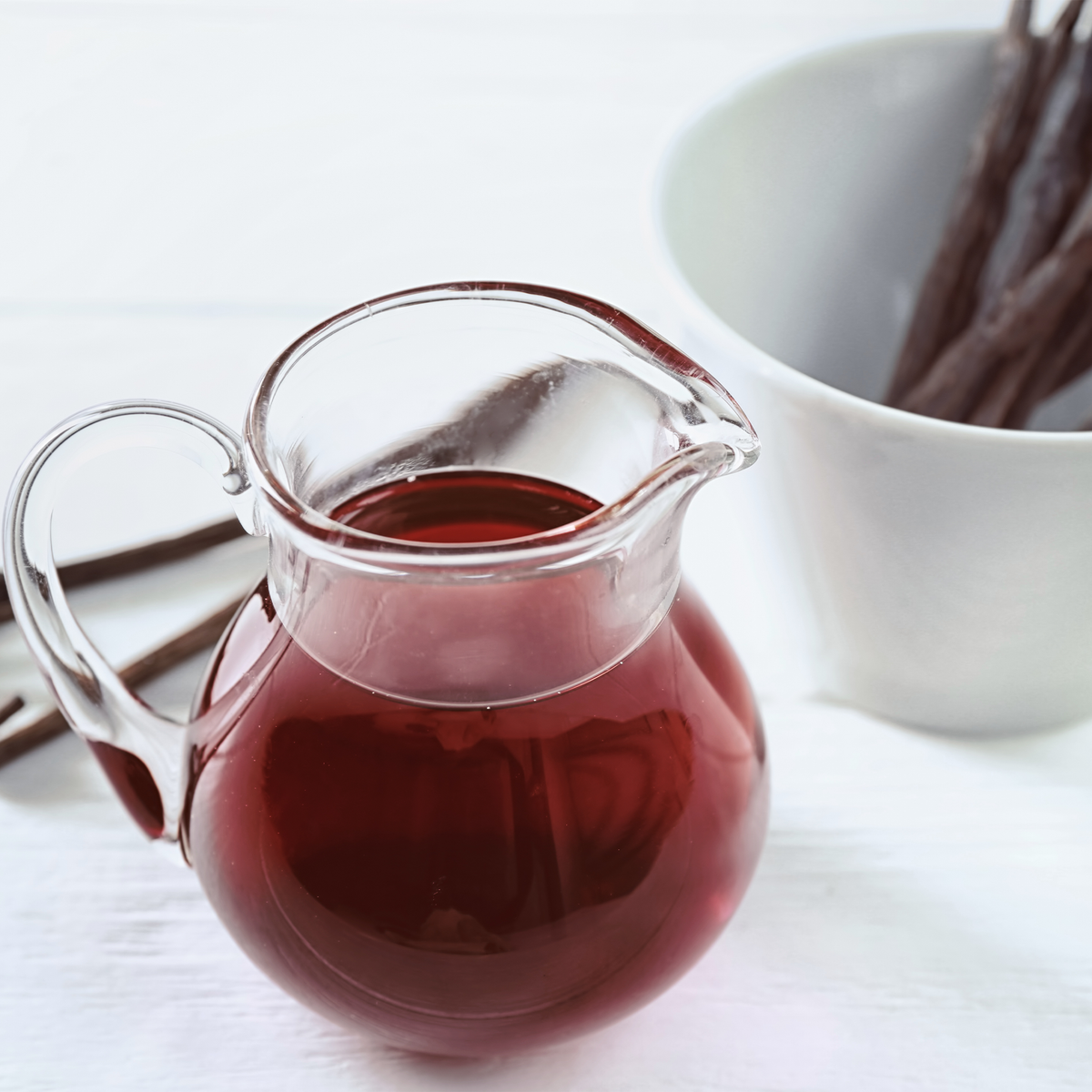 A clear glass pitcher filled with dark red liquid, set on a white surface with vanilla beans in a white cup in the background.