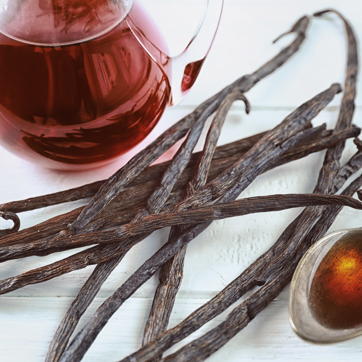 Vanilla beans spread across a white surface with a glass pitcher of amber tea and a small dish in the background.