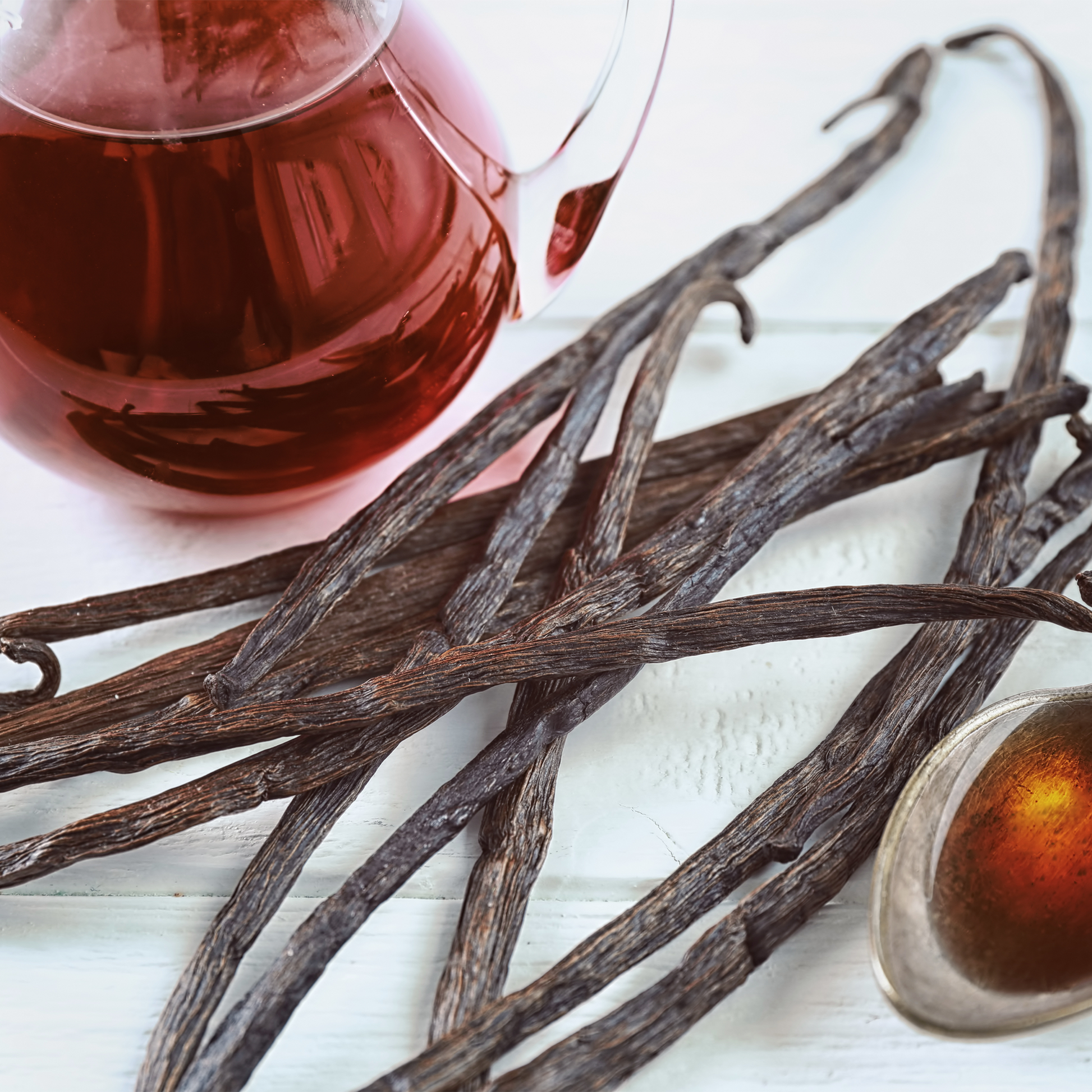 Vanilla beans spread across a white surface with a glass pitcher of amber tea and a small dish in the background.