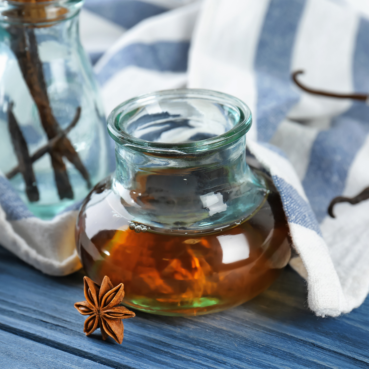 A small clear glass jar filled with vanilla extract styled on a blue wooden surface with a white and blue striped cloth, vanilla beans, and a star anise.