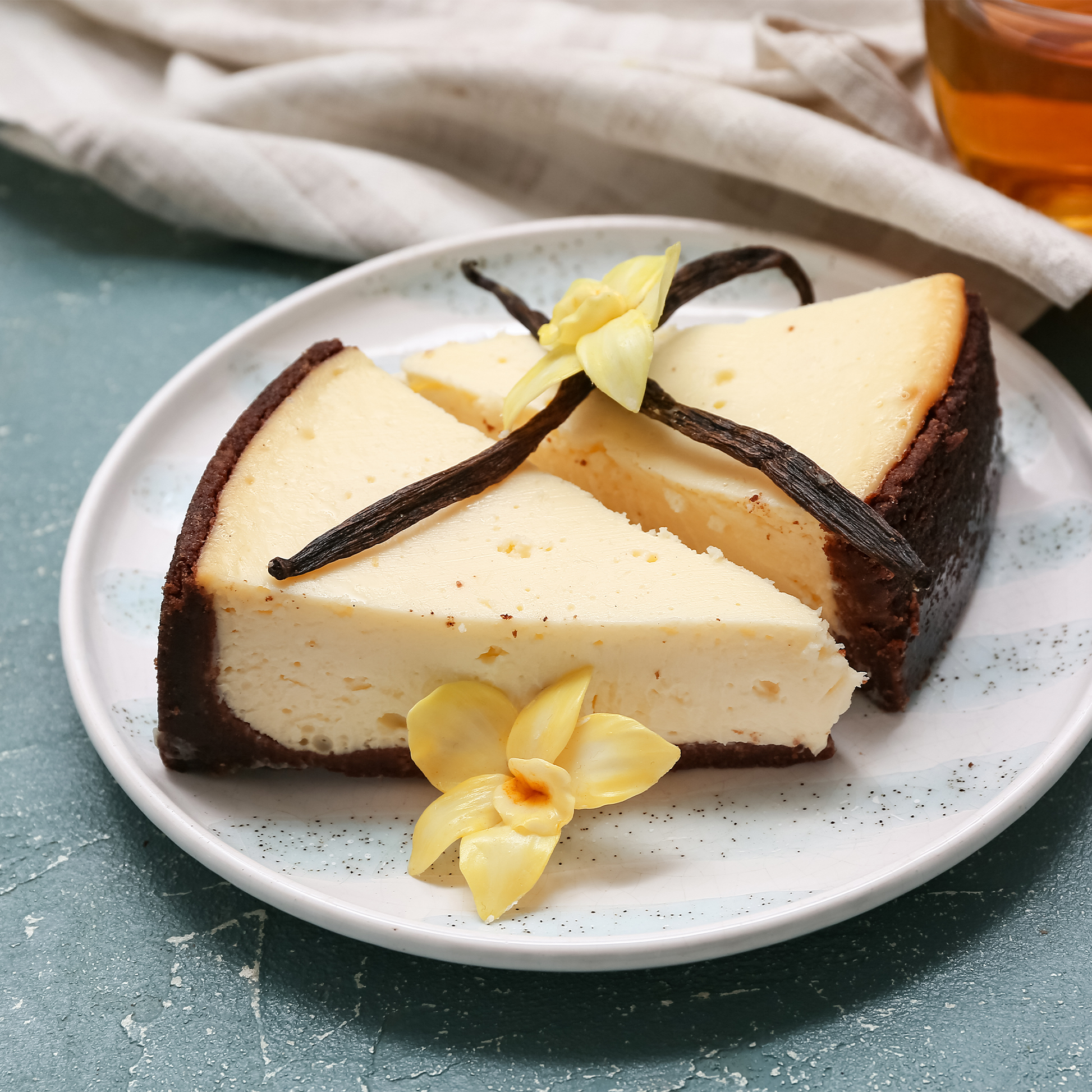 Two slices of cheesecake on a speckled white plate, garnished with vanilla beans and yellow flowers, set on a textured teal surface with a neutral cloth and amber drink in the background.