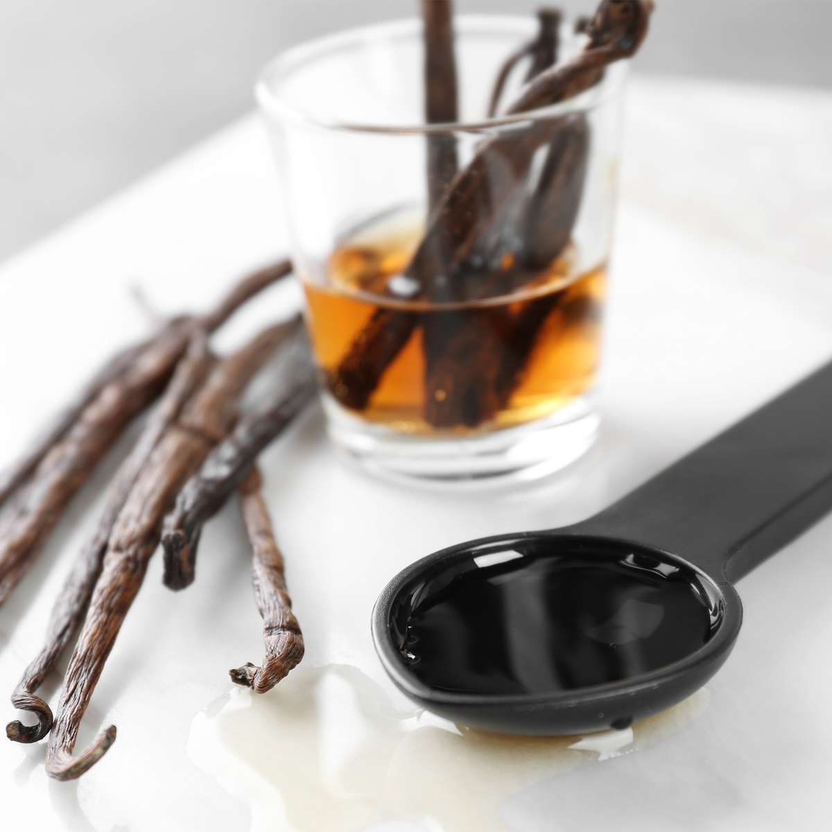 Vanilla bean pods in a clear glass with amber liquid, a black spoon filled with dark vanilla extract, and several vanilla beans on a white surface.