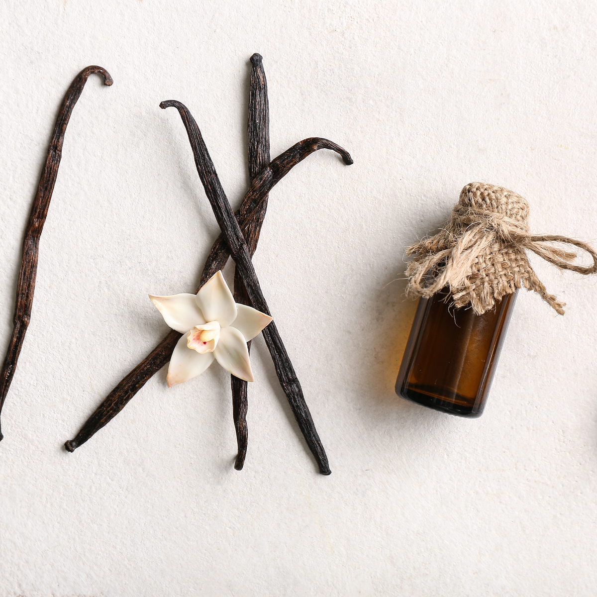An amber glass bottle with a rustic jute-wrapped neck and twine bow, arranged on a light textured background with vanilla beans, a vanilla flower, and a small white orchid.
