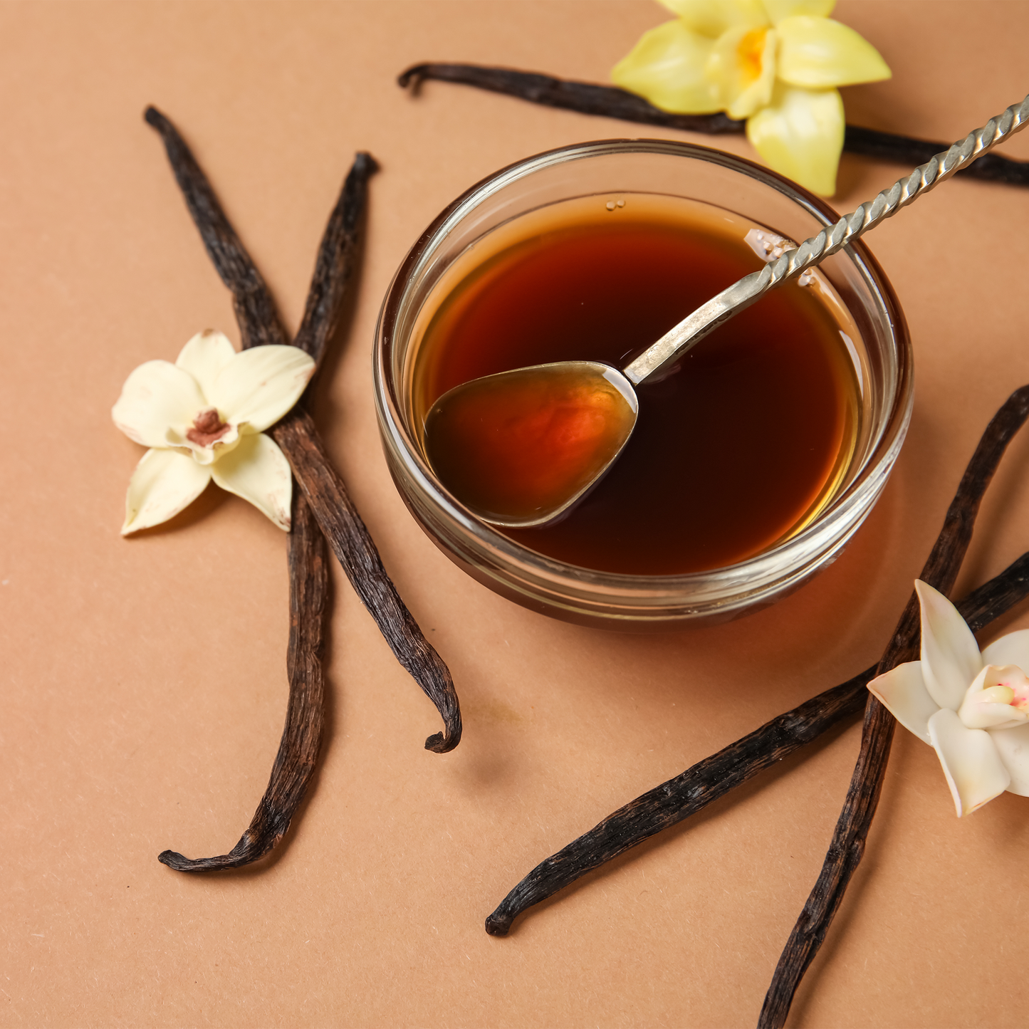 A small clear glass bowl filled with vanilla extract, with a metal spoon resting inside, styled on a warm tan background with vanilla beans and white orchid flowers.