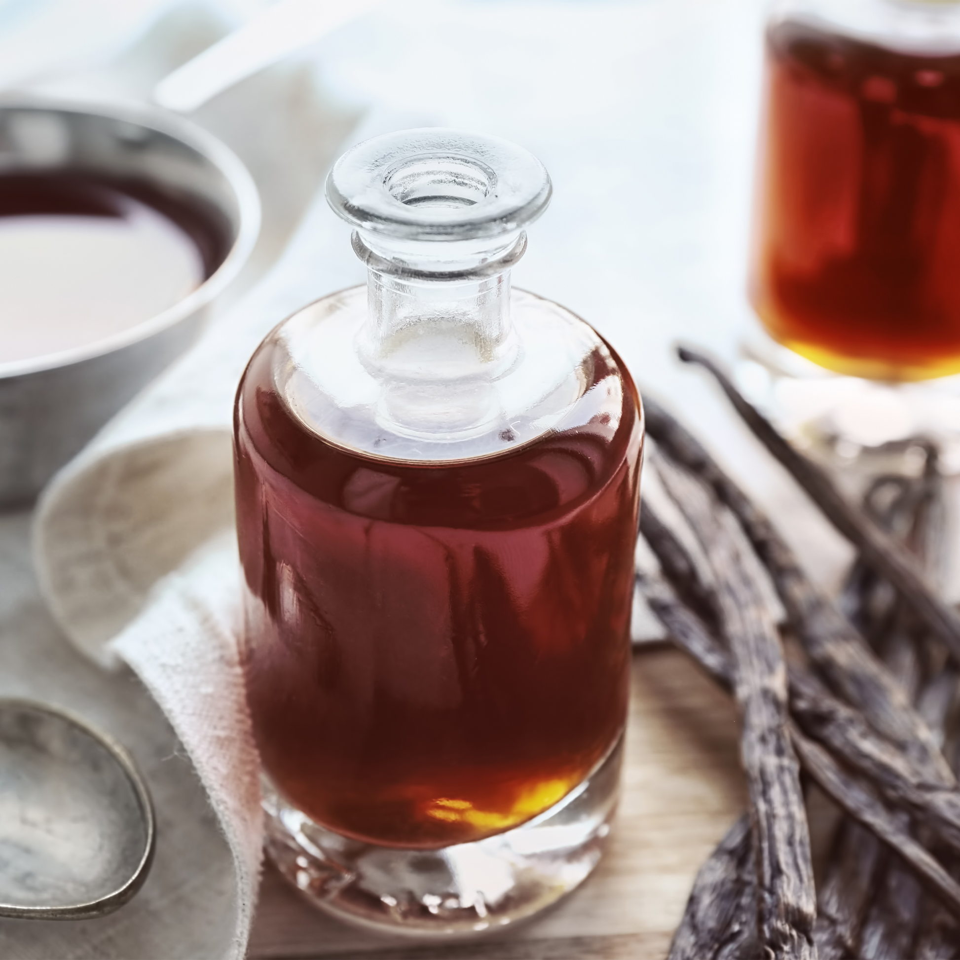 A clear glass decanter filled with vanilla extract, styled on a light surface with vanilla beans, a metal cup, and a blurred second decanter in the background.