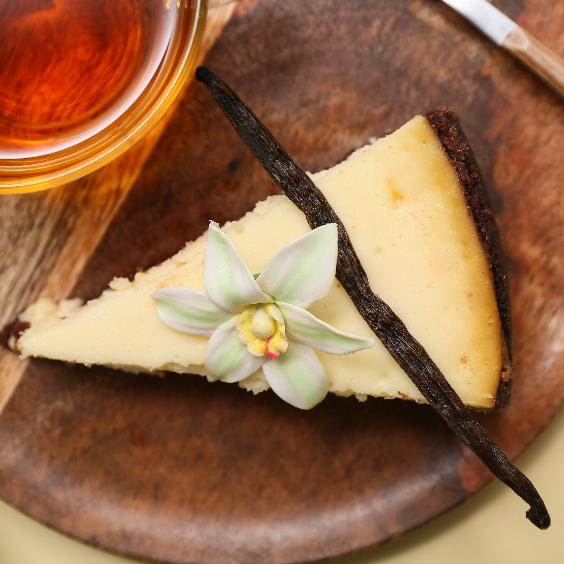 A slice of cheesecake on a rustic wooden plate, garnished with a vanilla bean and a small white orchid flower, with a glass of iced tea partially visible in the corner.