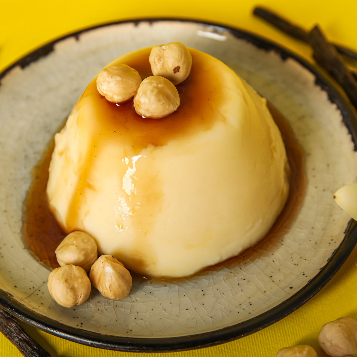 A close-up of a smooth, dome-shaped vanilla custard or flan dessert topped with caramel syrup and a few hazelnuts, served on a speckled ceramic plate against a bright yellow background.